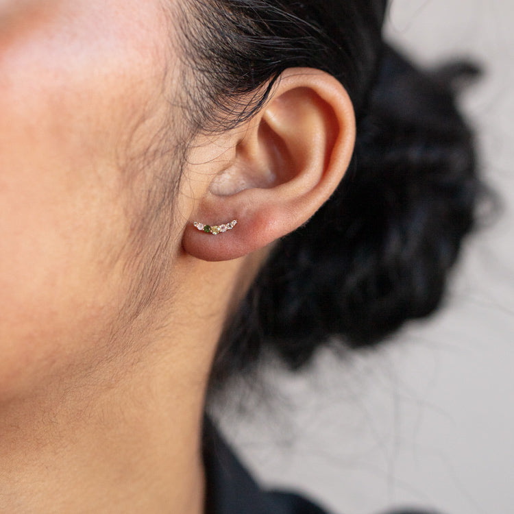 Close-up of a woman's ear adorned with Solid Gold Amour Acrostic Earrings featuring small gemstones, her dark hair in a loose bun and natural complexion. A solid gold necklace complements the minimal, neutral background.