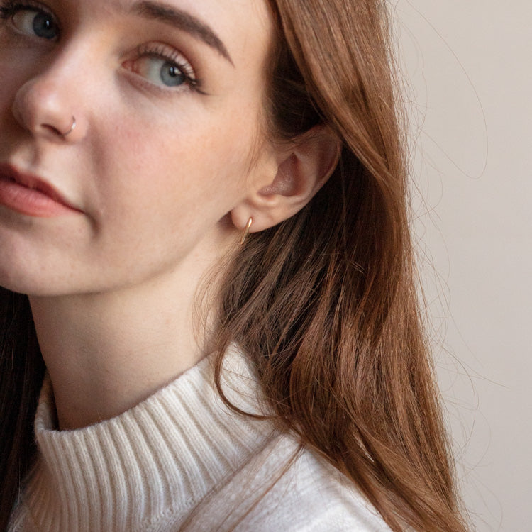 A young woman with long brown hair and light skin wears a cream turtleneck sweater and the Solid Gold Simple Oblong Hoops Sample, with a neutral background behind her.