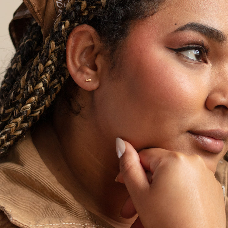 Close-up of a woman with braided hair, wearing a brown bandana and tan top, resting her chin on her hand. She has flawless makeup, neutral nails, and wears Solid Gold Dainty Birthstone Bar Studs for a refined touch.