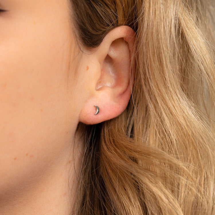 Close-up of a womans ear wearing Solid Gold Moon Earrings—a dainty crescent moon stud. She has light skin and blonde hair softly framing her ear.