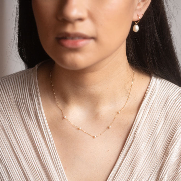 A woman, shown from the shoulders up against a neutral background, wears a cream textured V-neck top, a Solid Gold Delicate Pearl Chain Necklace, and a gold earring with a dangling pearl.