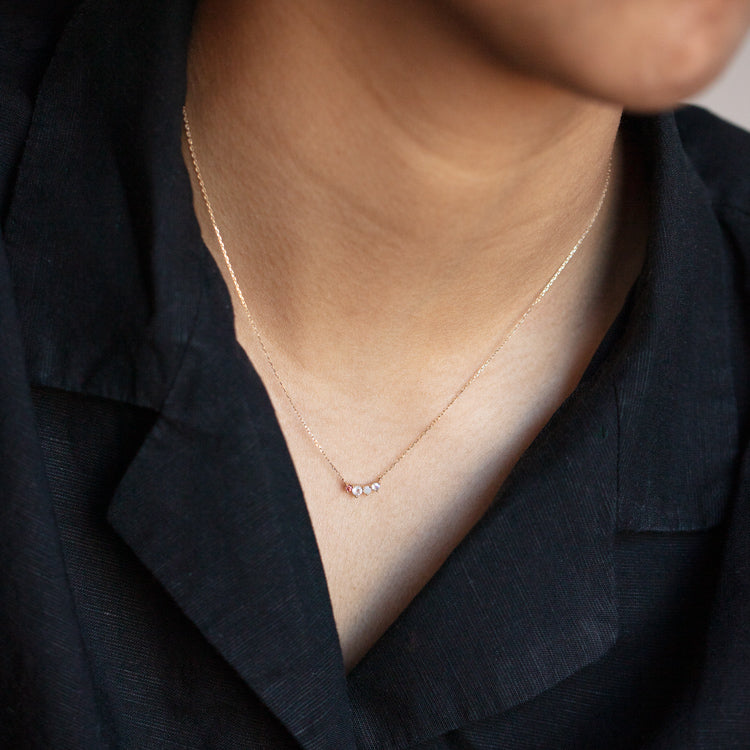 A close-up of a person wearing the Solid Gold Grow Acrostic Necklace Sample, a delicate 9k solid yellow gold chain with small beads, paired with a black collared shirt. The focus is on the necklace and neckline.
