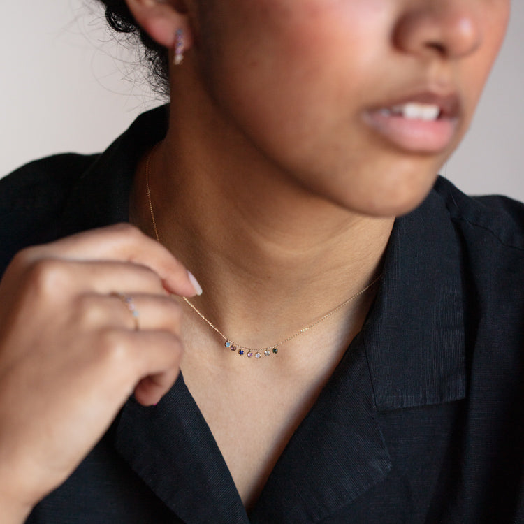 A person in a black collared shirt showcases the Solid Gold Balance Acrostic Necklace Sample—delicate 9k yellow gold with amethyst and blue calcedony charms—paired with a matching stud earring, focusing on the jewelry around the neck and ear.