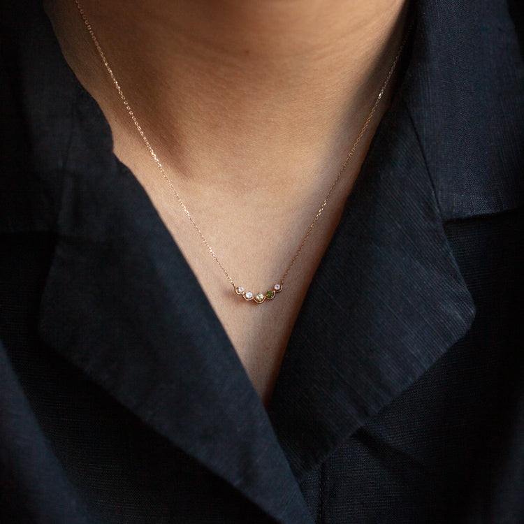 A close-up of a person wearing the Solid Gold Amour Acrostic Necklace Sample— a delicate 9k yellow gold chain with five colorful gemstones like rose quartz and aquamarine— against their skin and a dark collared shirt.