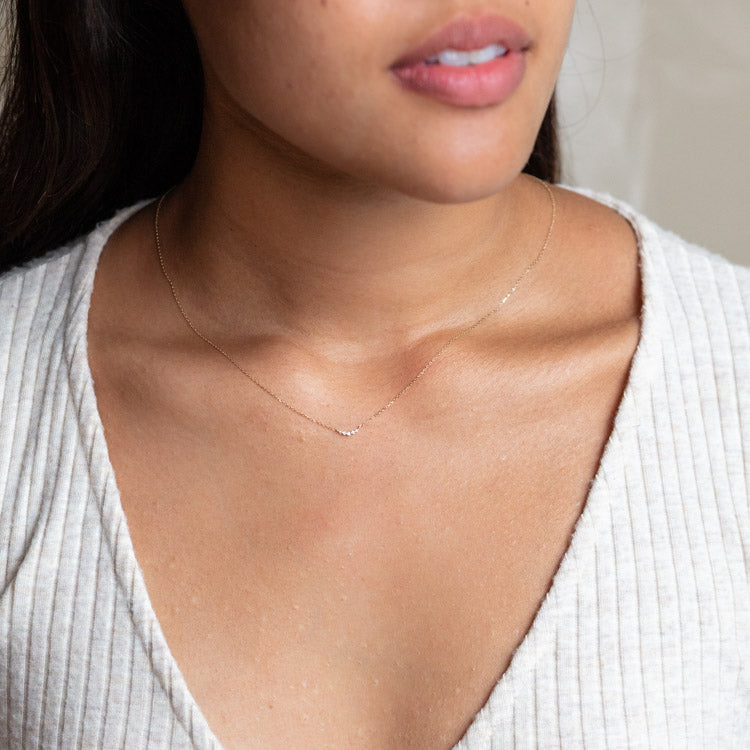 A close-up of a woman wearing the Solid Gold Diamond Arc Necklace Sample, a delicate 9k yellow gold chain with a subtle diamond arc pendant. She pairs it with a textured cream v-neck top, highlighting the elegant 16-inch necklace on her collarbone.