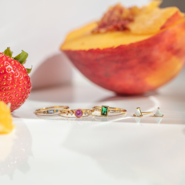 The Iridescent Opal Triangle Studs and a 14k yellow gold plated bracelet with colorful gemstones are displayed on a white surface, with a strawberry and sliced peach in the background.