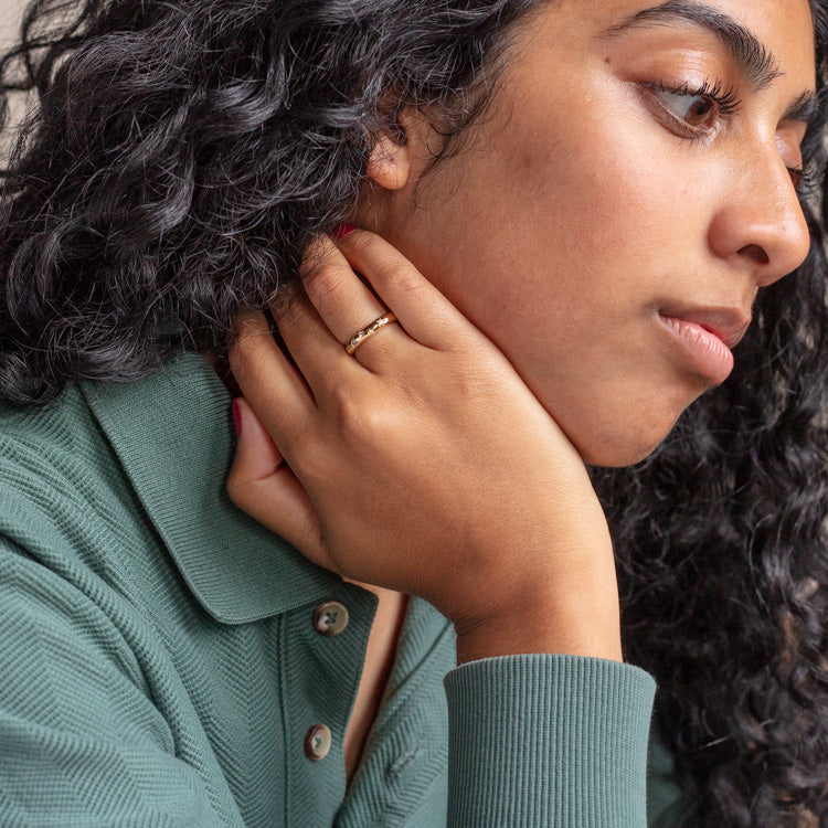 A person with long, curly black hair rests their hand on their neck, showing off the Star Crossed Celestial Dome Ring. They wear a green collared shirt and gaze thoughtfully to the side.