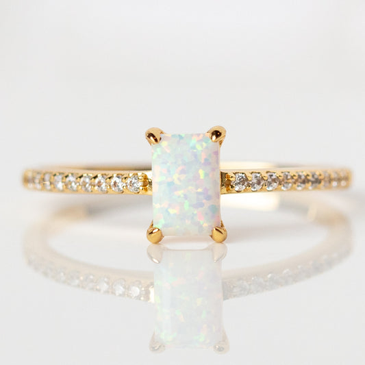 The Hopeful Opal Baguette Ring showcases a rectangular opal center with iridescent hues, set in yellow gold and accented by a pave band of clear cubic zirconia, beautifully displayed on a reflective white surface.