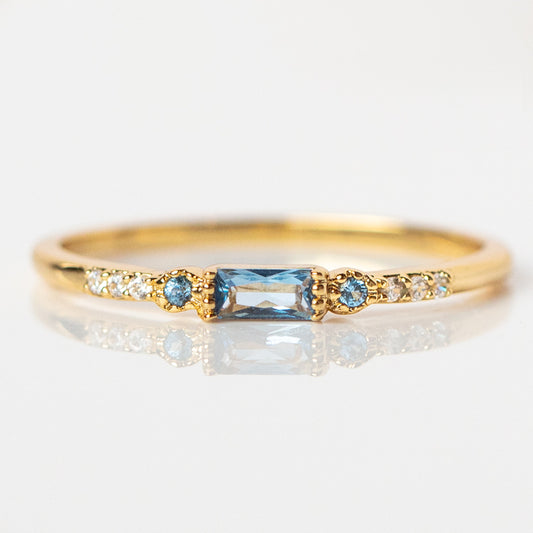 The Calming Blue Stacking Ring showcases a rectangular blue CZ center stone, accented by small round blue stones and tiny clear stones along the delicate gold band.