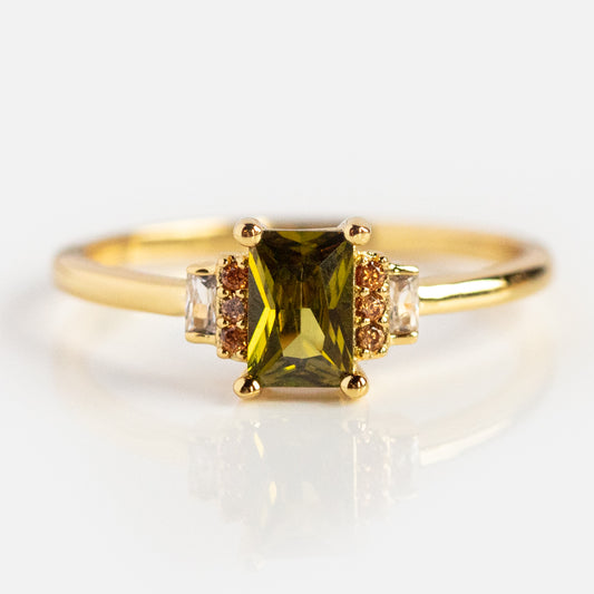 The Retro Era Ring features a rectangular green cubic zirconia centerpiece, two small clear stones, and two tiny orange stones set in gold, shown on a white background.