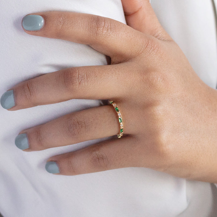 A hand with neatly manicured, light gray nails rests on white fabric. The ring finger features the Gatsby Stacking Ring, a delicate 14k yellow gold plated band set with small, green rectangular gemstones.