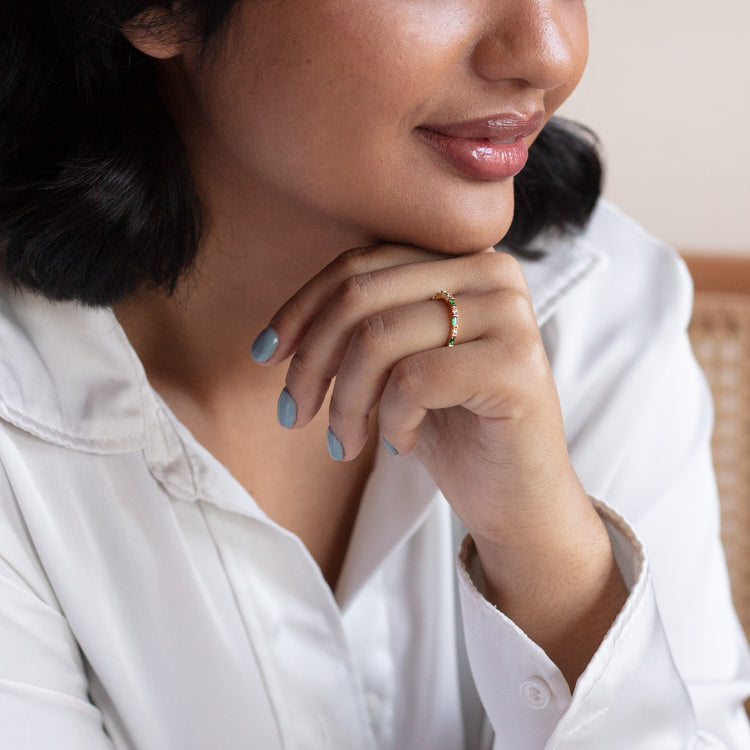 A woman in a white collared shirt smiles softly, showing off light blue nails and the Gatsby Stacking Ring—an elegant green CZ piece plated in 14k yellow gold. Her dark hair is styled in a bob as she rests her chin on her hand.