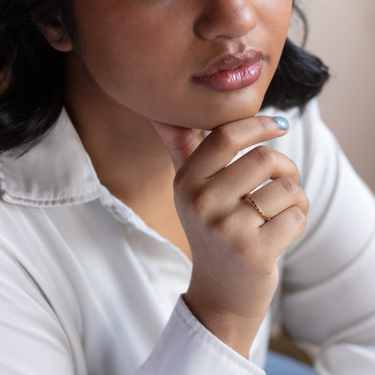 A person in a white collared shirt rests their chin on their hand, displaying the Regency Stacking Ring—a delicate yellow gold plated band—on their finger, with light blue nail polish and slightly parted lips.