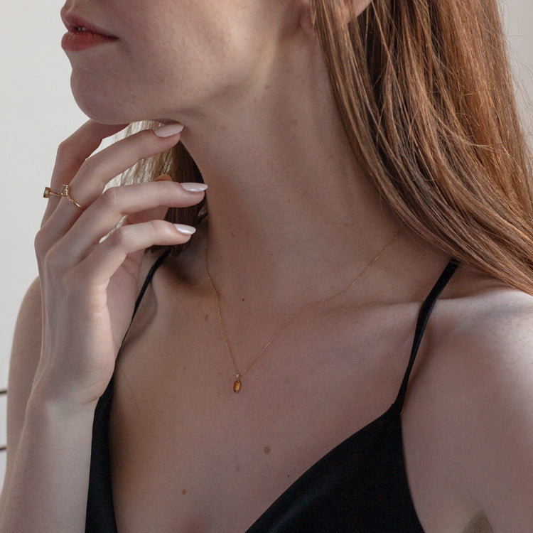 A woman with long, light brown hair models the Solid Gold Citrine Destiny Hexagon Necklace for Abundance (16", 9k yellow gold) with a small citrine pendant, a black spaghetti strap top, a gold ring, and a natural manicure.