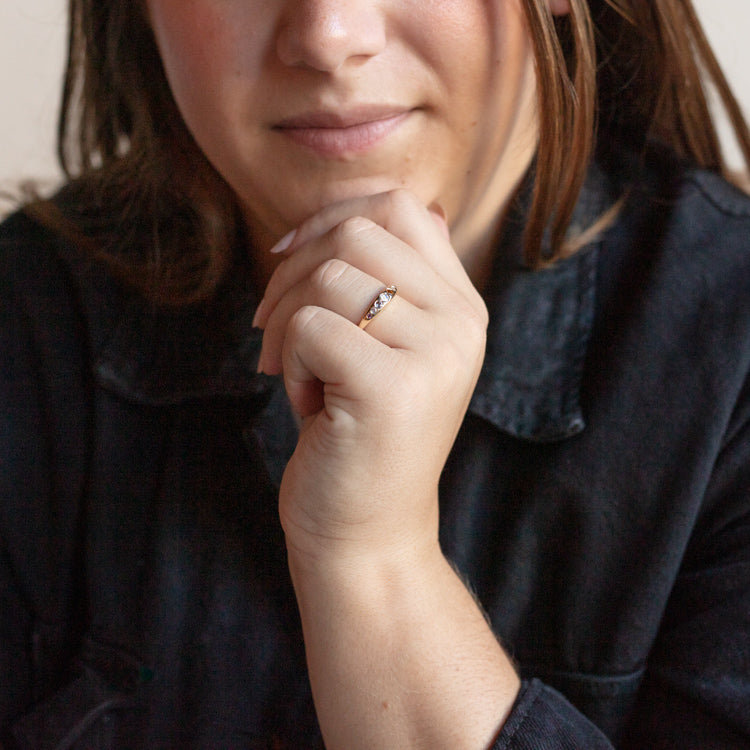 A person in a dark shirt rests their chin on their fist, showcasing the Shes an Artist Ring in Yellow Gold by I Like it Here Club. Their face is partly visible, and their brown hair falls loosely around them.