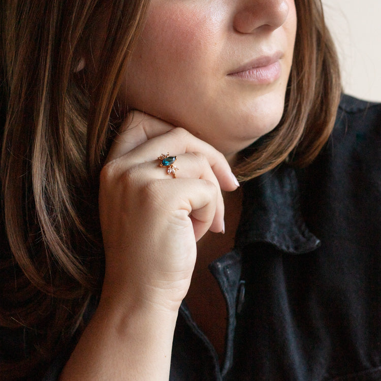 A woman with light brown hair, wearing a dark top, rests her chin on her hand and showcases the Gemstone Firefly Ring—yellow gold vermeil with colorful gemstones and a flower charm—while looking thoughtful.
