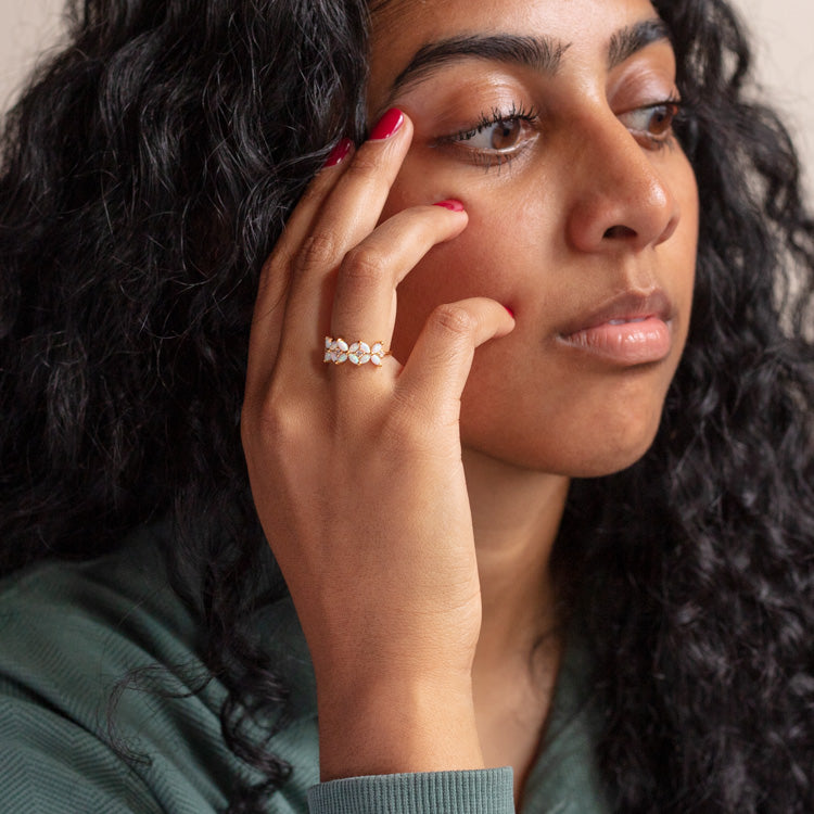 A woman with long curly black hair touches her face, showcasing the Opal Fae Ring—a yellow gold floral design—on her finger. She has natural makeup, red nail polish, and wears a green top, appearing thoughtful.