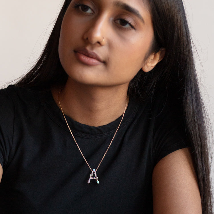 A woman with long dark hair, in a black shirt, looks to the side. She wears an Opal Monogram Pendant necklace featuring a gold letter A.