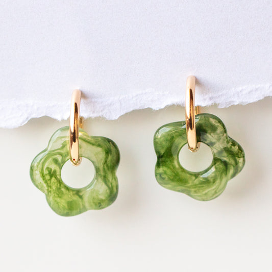 The Goodies Hoop Earrings, featuring yellow gold plated hoops and green marbled flower pendants, are displayed against a white background with a torn paper edge at the top.