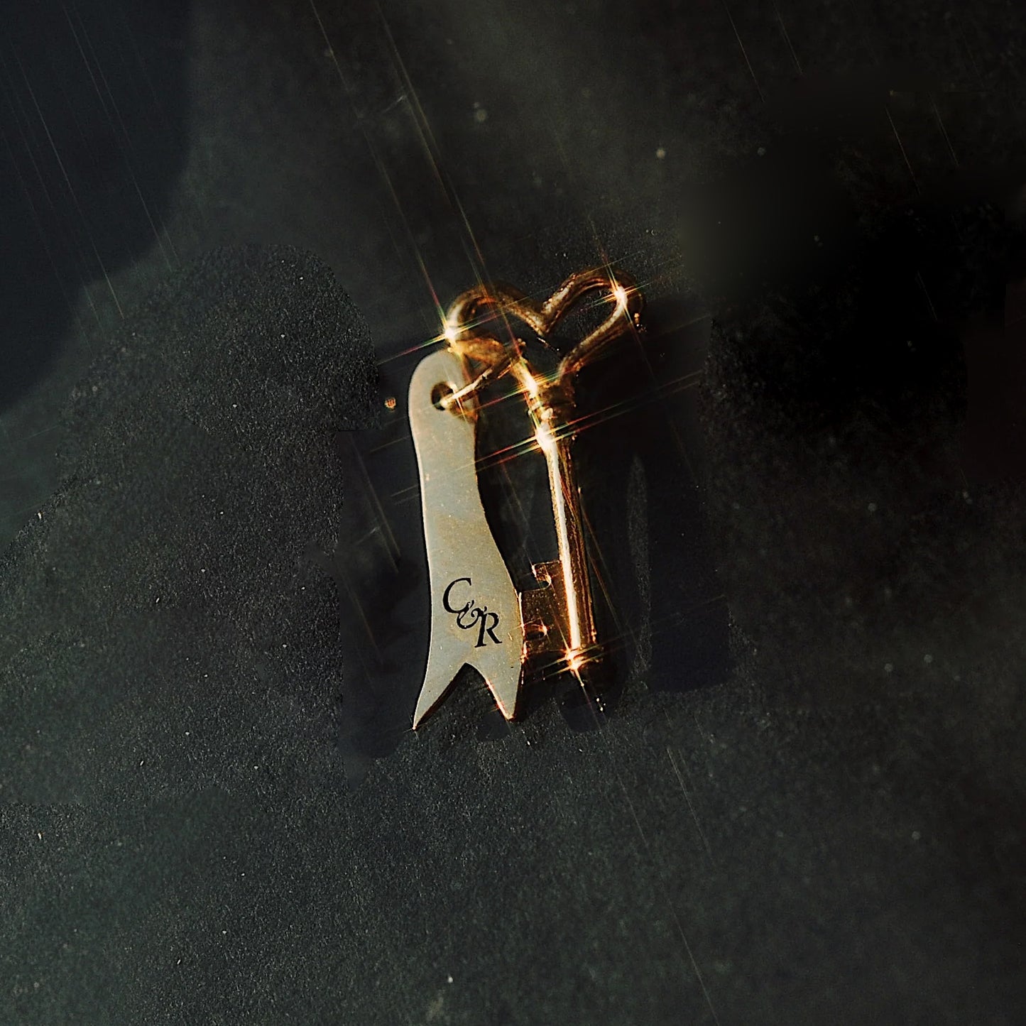 The Customizable You and I Necklace features a gold engraved key and a silver ribbon-shaped COR charm, displayed on a dark textured surface under soft lighting—an elegant, personalized jewelry piece.