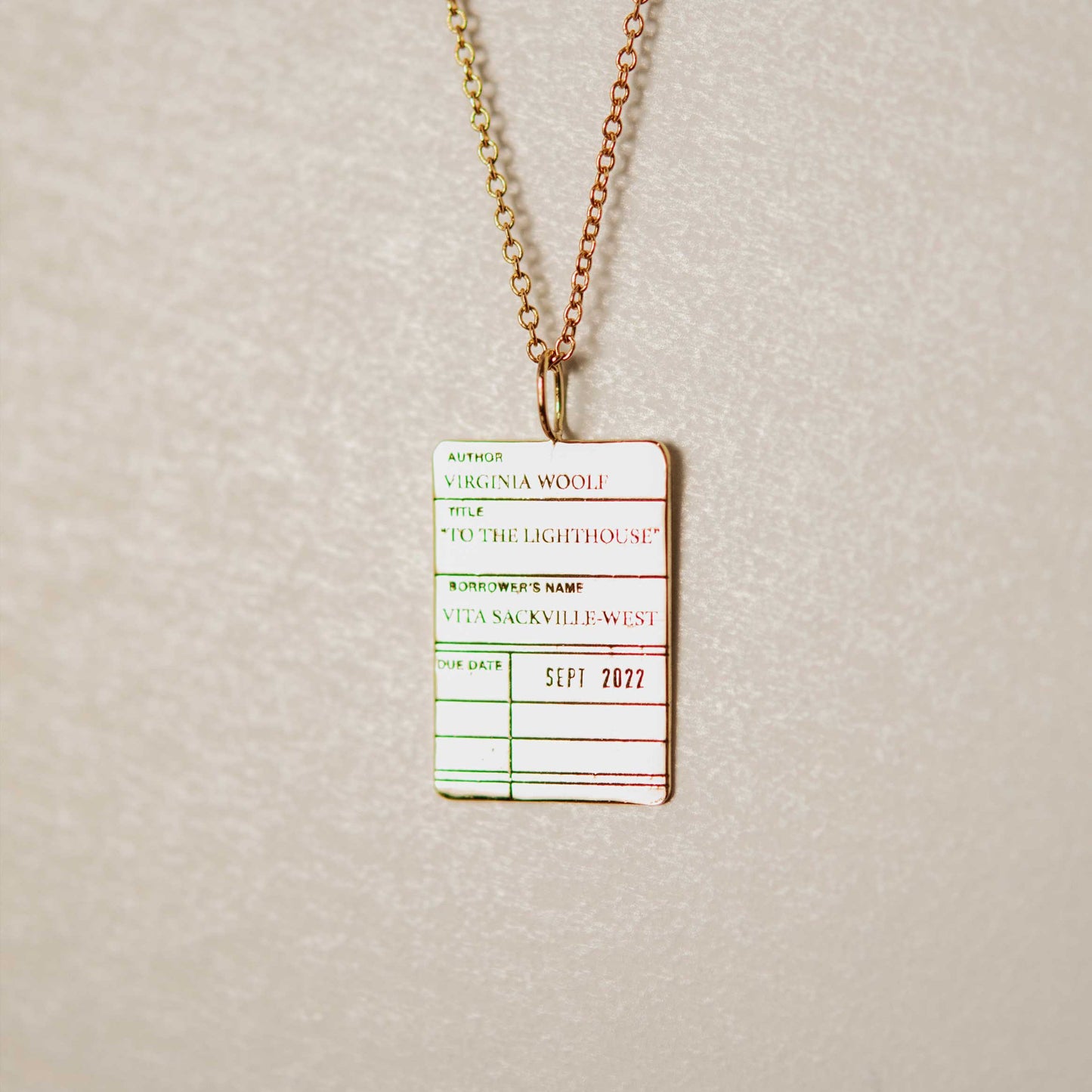 The Customizable Library Card Necklace features an engraved gold pendant shaped like a vintage card for Virginia Woolf’s To the Lighthouse, with borrower Vita Sackville-West and due date Sept 2022, on a light background.