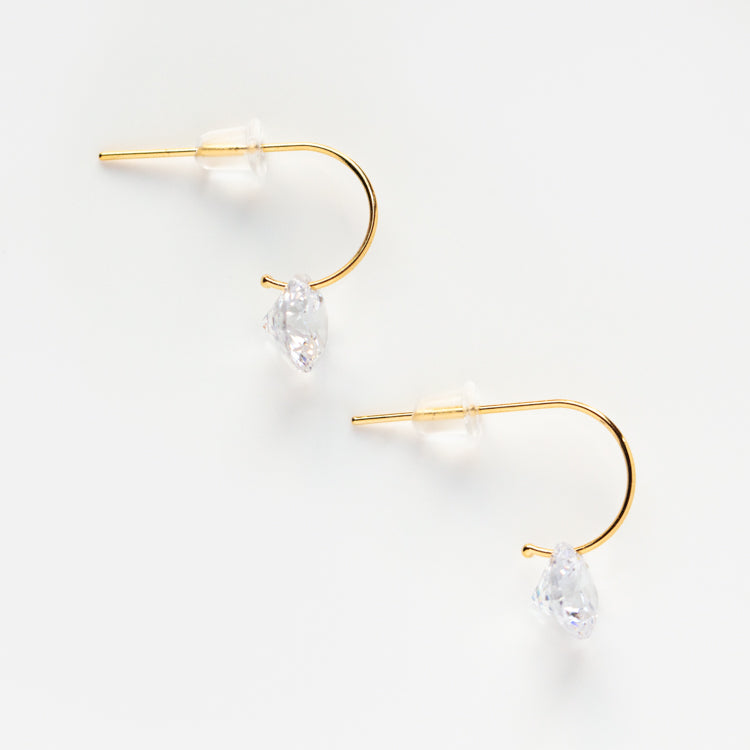 The Floating Solitaire CZ Huggie Earrings, gold with clear plastic backs and delicate teardrop-shaped CZ drops on curved wires, are displayed on a white background.