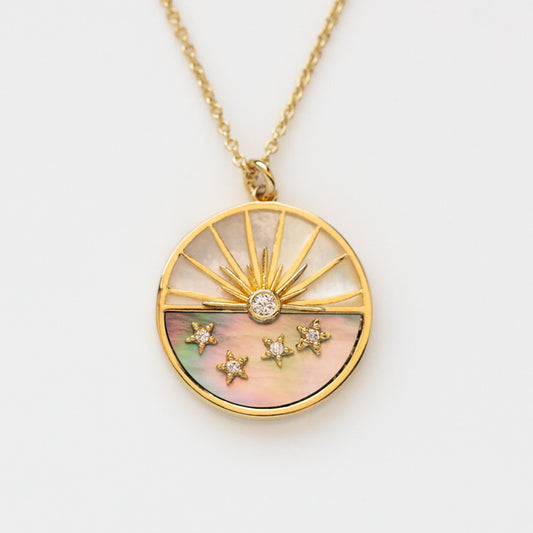 The Abalone and Mother of Pearl Horizon Necklace features a round gold pendant with a sunburst design, central crystal, three star crystals, and a shimmering abalone background on a chain.