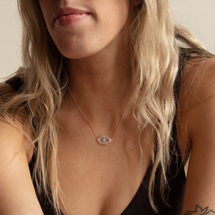 A woman with wavy blonde hair wears the CZ Evil Eye with Opal Center Necklace, featuring a central opal, paired with a black sleeveless top. Her lips and chin are visible, but the rest of her face is partially out of view.