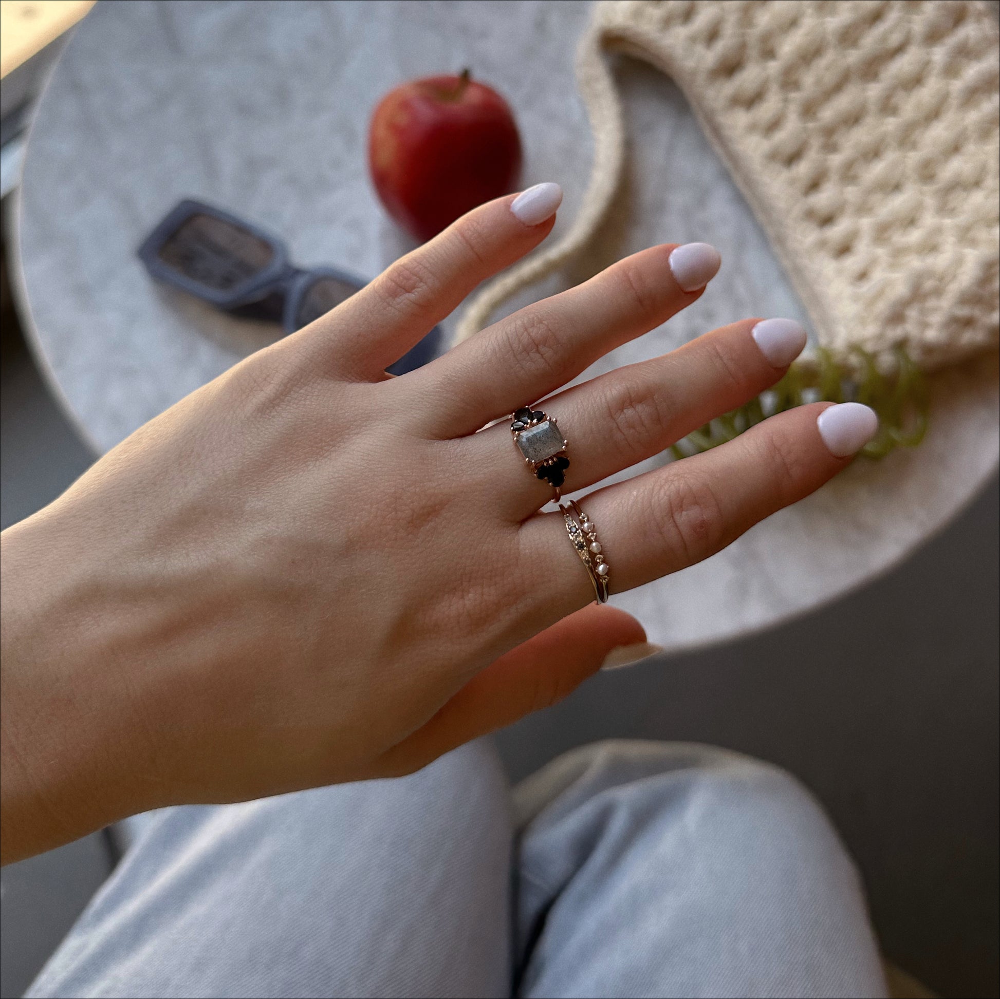 A hand with pale pink nails wearing the Gemstone Empress Ring and two others rests on a lap; in the background, a table displays an apple, dark sunglasses, and a textured beige bag.