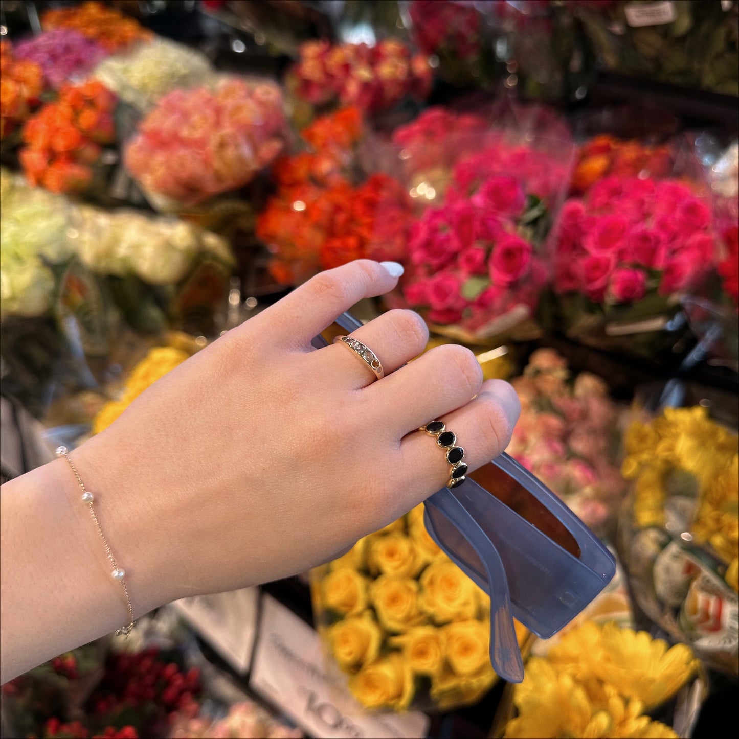 A hand adorned with rings—including the Shes an Artist Ring in Yellow Gold by I Like it Here Club—holds blue sunglasses before vibrant rose bouquets in a flower shop.