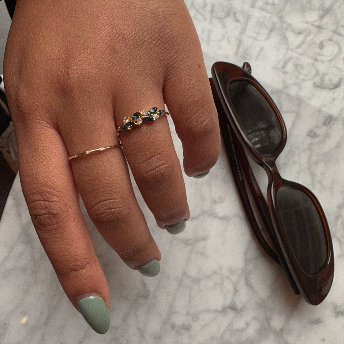 A hand with sage green nail polish wears two rings—a thin gold band and the Gemstone Orchard Ring—resting on a marble surface beside dark brown sunglasses.