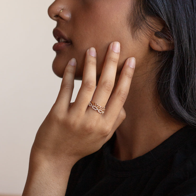A person with medium skin tone rests their hand on their cheek, showcasing the Solid Gold White Sapphire Chunky Chain Link Ring on their finger. They have long dark hair, a nose stud, and wear a black shirt.