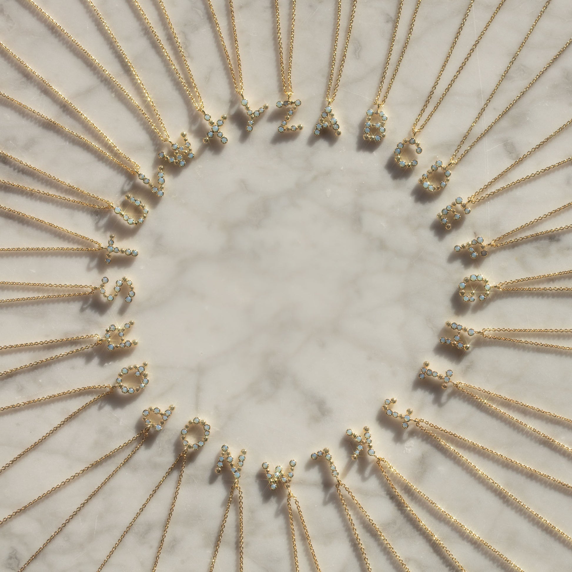 Initial Charm Necklace Sample features brass-based, 14k yellow gold-plated necklaces with opalescent nano gem letter pendants, arranged in a circle on marble and evenly spaced.