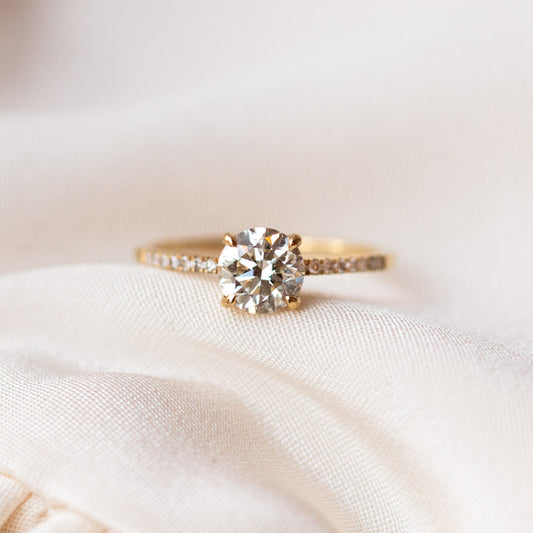 The 9kt Round Cut Diamond Solitaire Engagement Ring with Pave Diamond Band (Sample Size 7) is displayed on a soft, cream-colored fabric background.