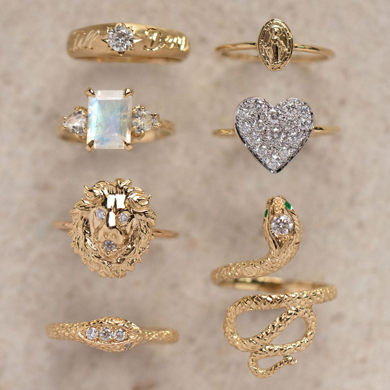 Eight ornate 14k yellow gold rings are arranged vertically on a beige surface, featuring designs including the Empress Moonstone Ring, a white sapphire ring, lion head, snake, sparkling heart, and engraved or textured patterns.
