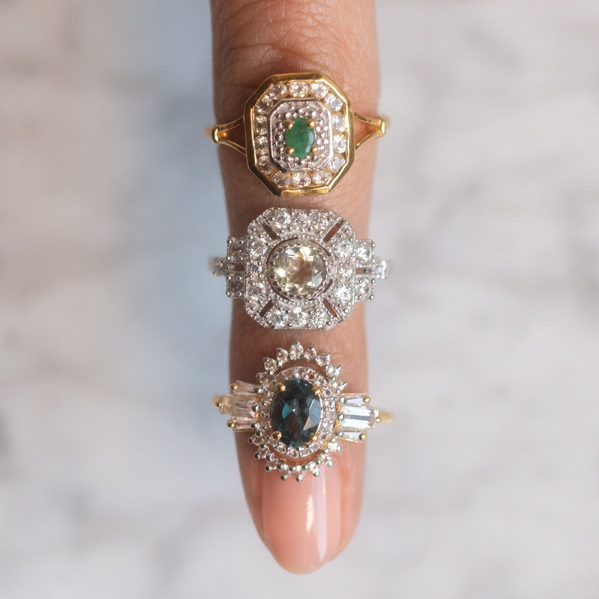 A close-up of a hand wearing the Venus Ring, a vintage-inspired piece with green amethyst and intricate gold and sterling silver settings, shown against a softly blurred background.