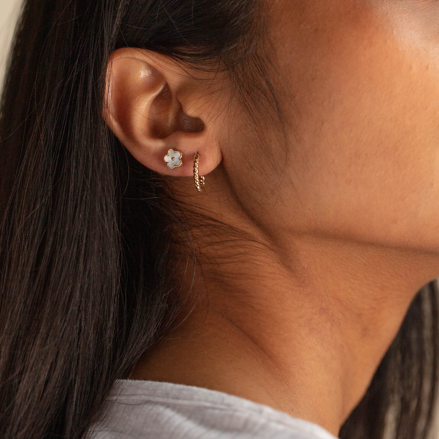 A close-up of a woman wearing Solid Gold Twisted Huggie Hoops and a small white flower stud. Her long dark hair is tucked behind her ear, and she is dressed in a light-colored top.
