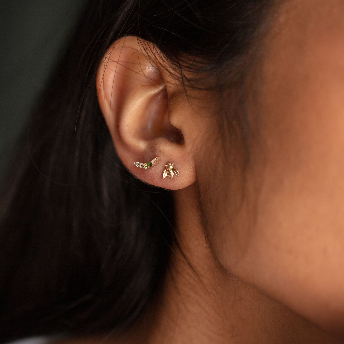 Close-up of a woman's ear wearing Golden Bee Earrings: 14k gold filled, featuring a delicate bumble bee stud and a small curved bar with green stones. Her dark hair is tucked behind her ear.