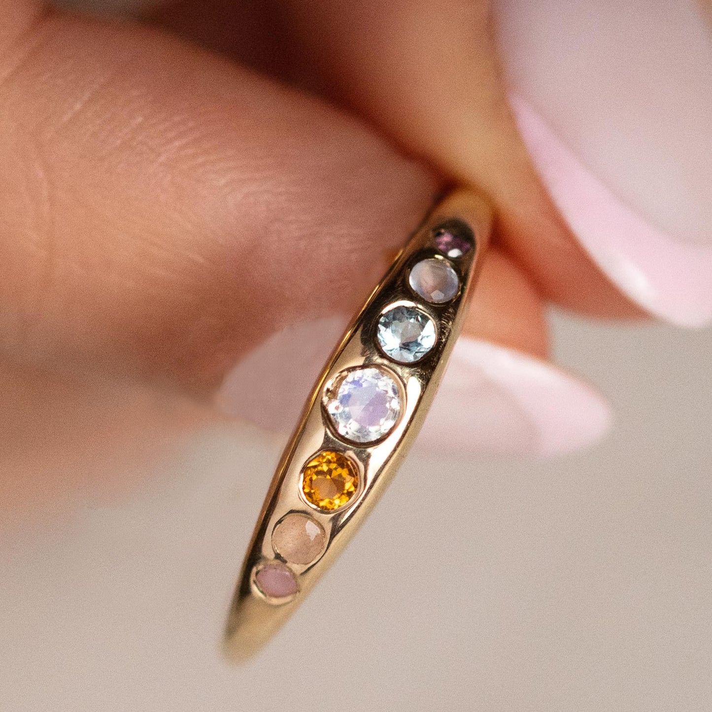 A close-up of a hand holding the She's an Artist Ring in Yellow Gold, featuring five vibrant gemstones—blue, pink, orange, and white—set in 10K gold against a softly blurred background.