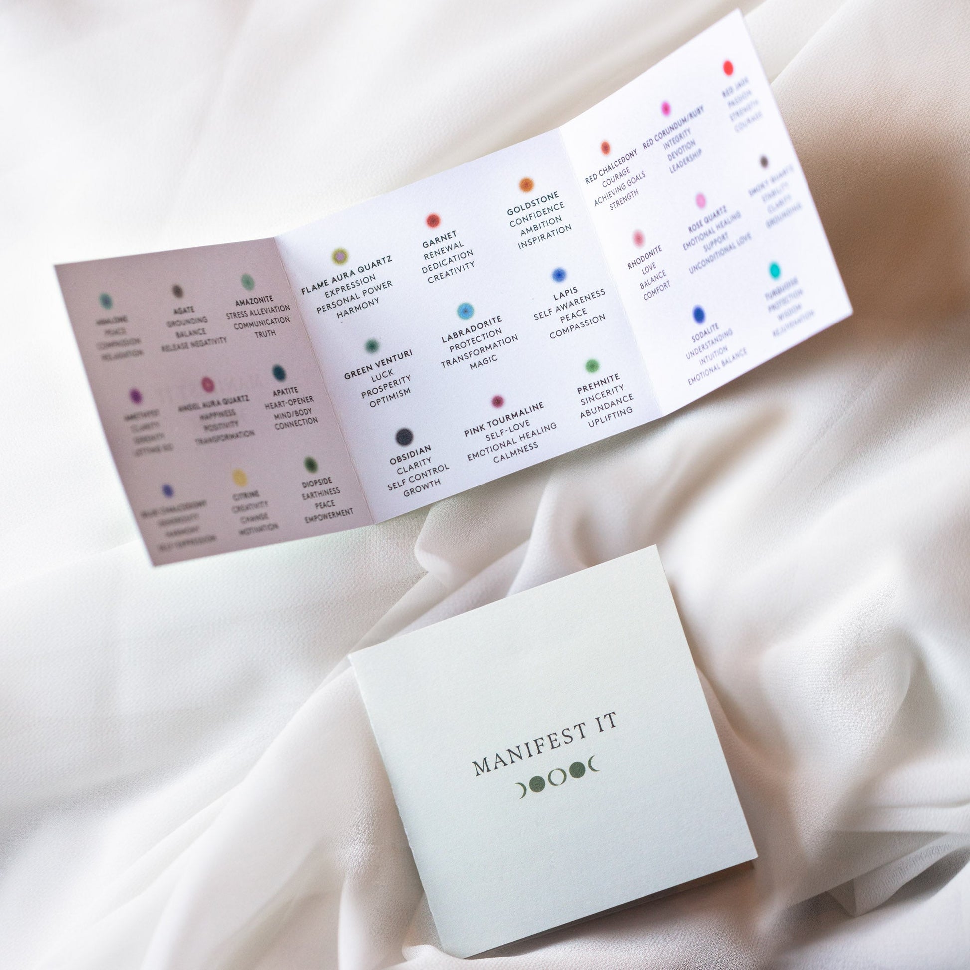 A folded card labeled MANIFEST IT rests on white fabric beside an open guide with colorful dots, crystal descriptions like iolite, and a Solid Gold Crystal Ring for Manifestation (Abalone, Sample Size 7) with their unique meanings.