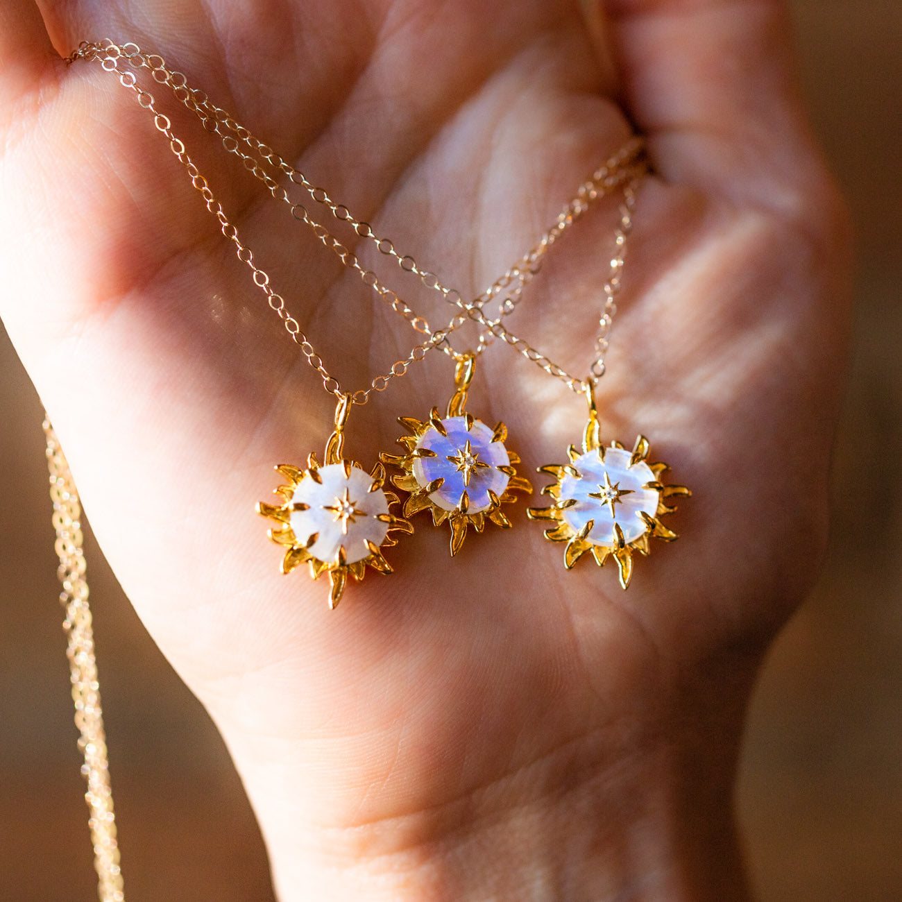 A hand holds three "You Are My Forever Sun, Moon & Star" pendants, featuring gold sunburst designs with iridescent moonstone accents, each hanging from a delicate gold chain.