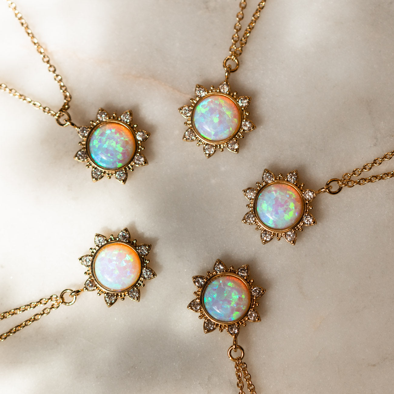 Four White Opal Sun Necklaces, each gold plated with a round white opal pendant and a sunburst halo of cubic zirconia, displayed on a light marble surface.