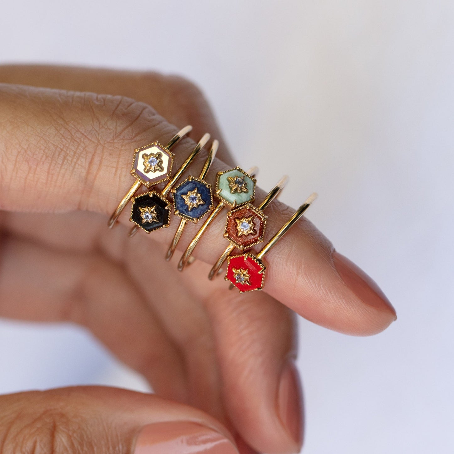 A manicured hand shows five 9k solid yellow gold rings. Each hexagonal face—in black, navy, light blue, red jade, or cream—shows a star design with a central gem. Shown: Solid Gold Crystal Ring for Manifestation Red Jade (Size 8).
