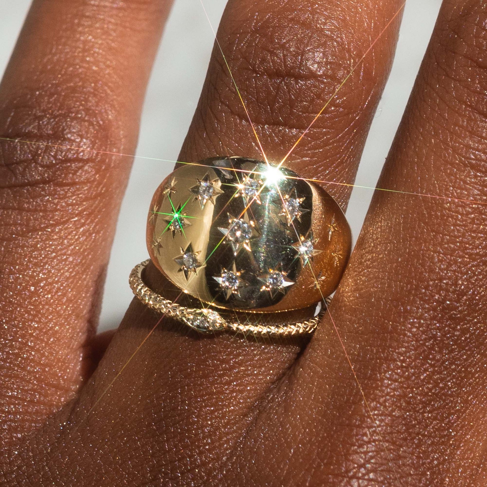 A close-up of a hand wearing two rings: the Ouroboros Diamond Snake Ring in shiny 14k solid gold with star-shaped diamonds, and a twisted gold band below, both glinting in bright light that creates a lens flare.