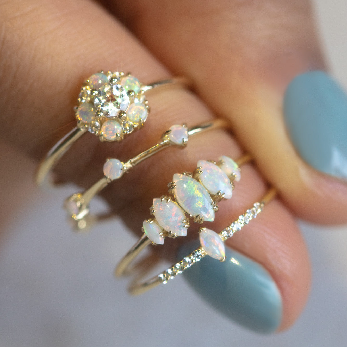 A hand with pale blue nail polish displays the Solid Gold Essential Opal Marquise Cluster Ring in size 7, highlighting its unique gold setting and shimmering opal and diamond cluster.