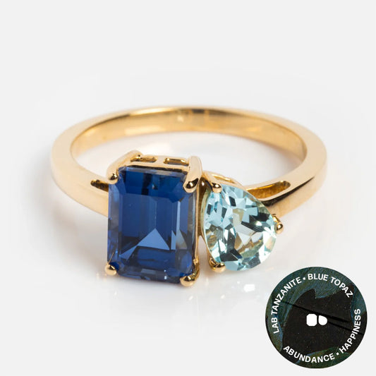 The Solid Gold Toi et Moi Colorful Gemstone Ring features a rectangular lab tanzanite and a pear-shaped blue topaz—unique gemstones symbolizing abundance and happiness, perfect for any occasion.