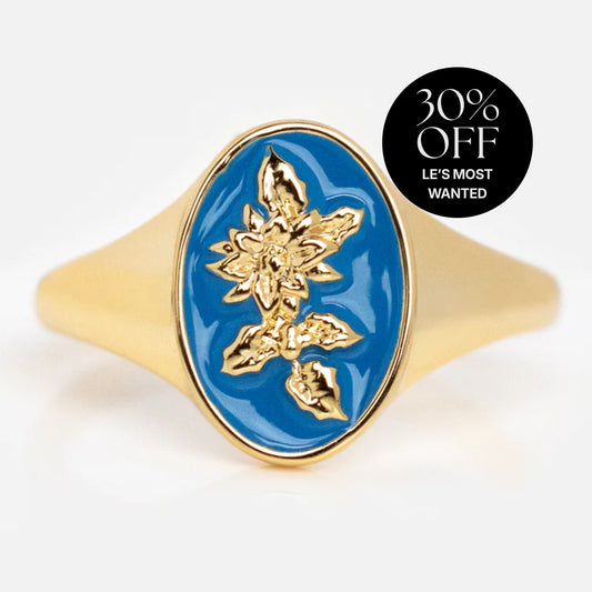 The Enamel Birth Flower Signet Ring is a 14k gold plated ring with a blue oval face and raised gold floral design. A black circular label in the top right says "30% OFF LES MOST WANTED." The background is plain white.