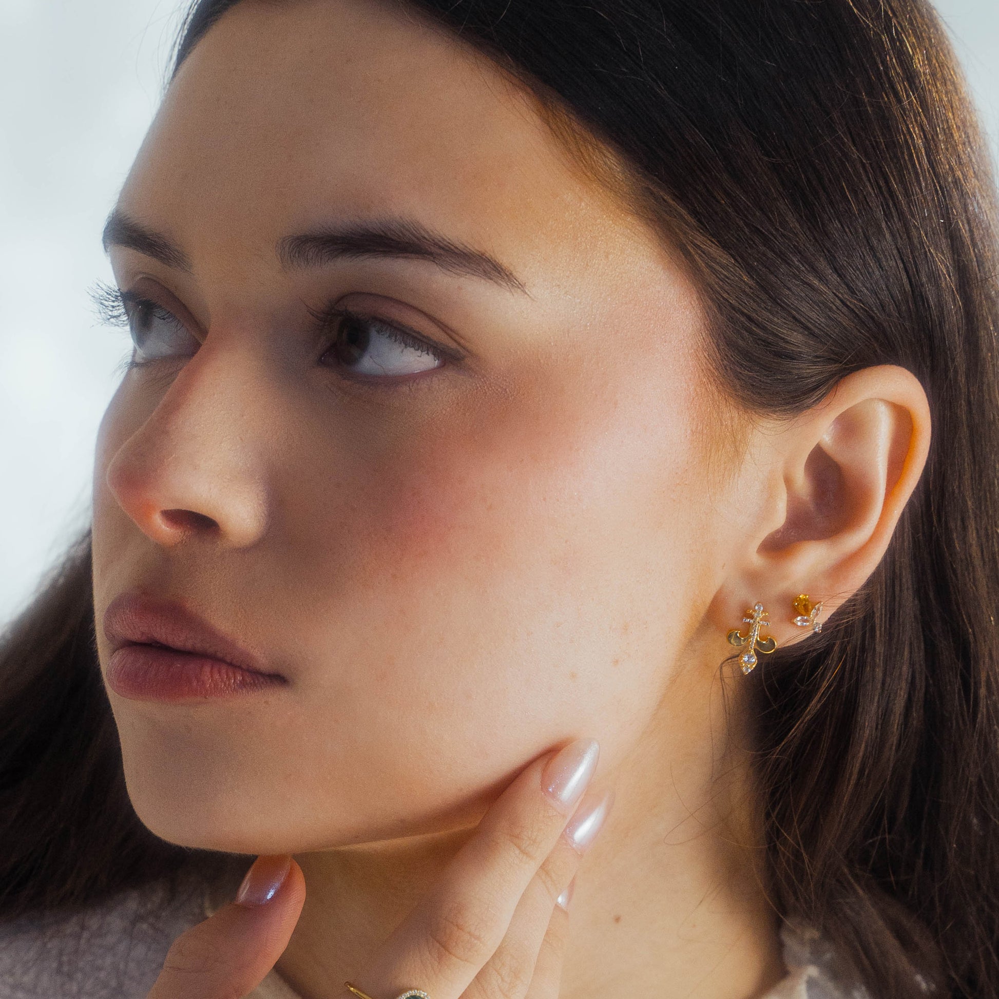 A woman with long dark hair touches her chin, wearing Jazz Age Citrine Studs. She has a calm, thoughtful expression as she gazes to the side; the softly blurred background enhances her elegant look.