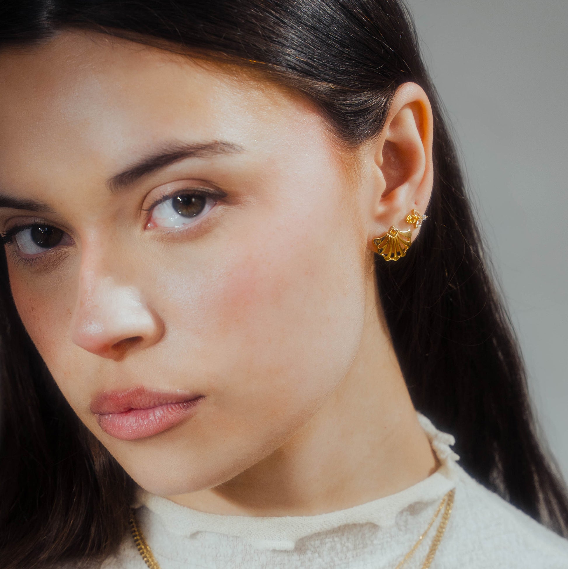 A woman with long dark hair and a neutral expression wears Jazz Age Citrine Studs in 14k yellow gold vermeil and layered gold necklaces, dressed in a cream-colored top with a ruffled collar.