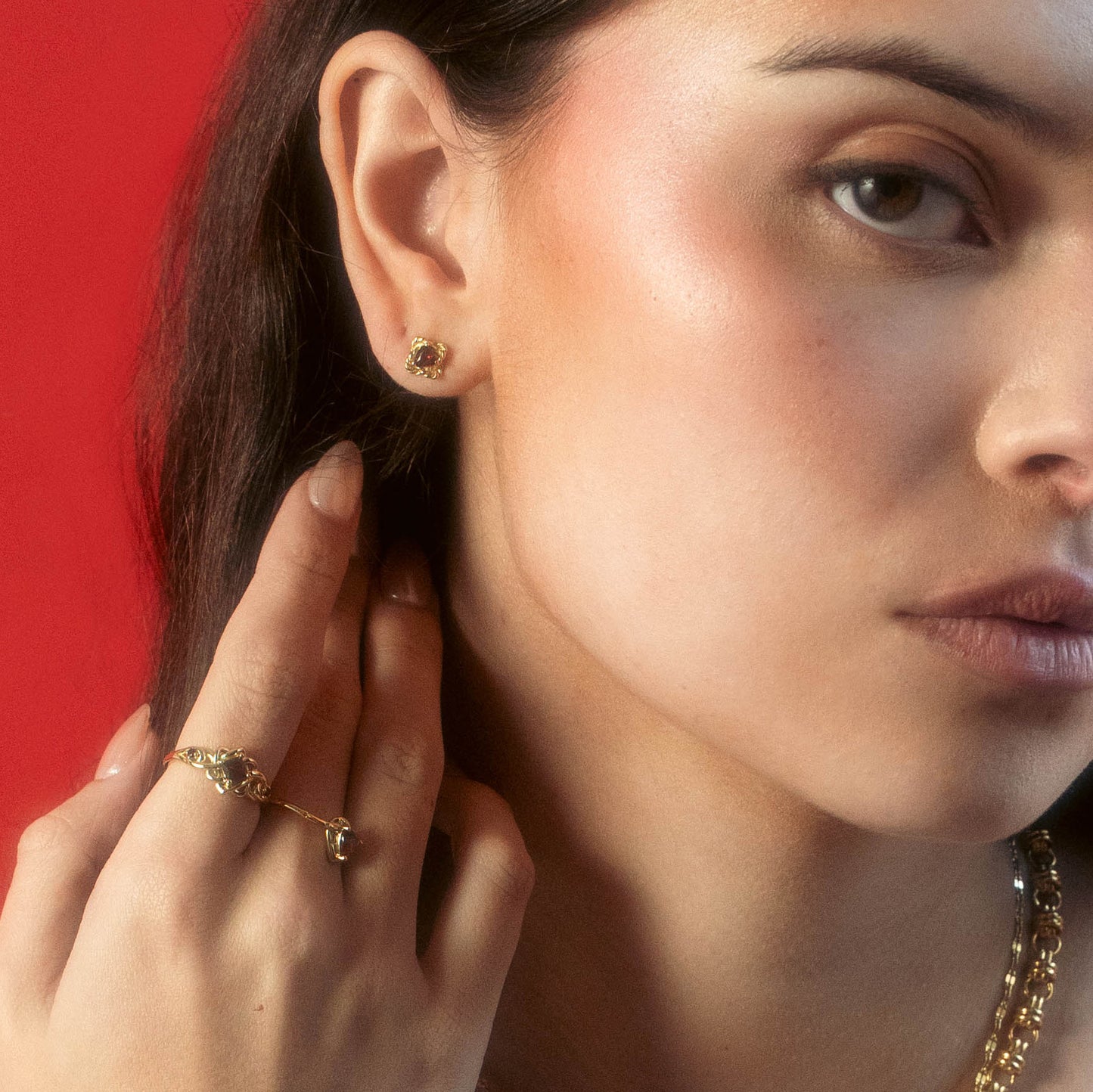 A woman with long dark hair poses against a red background, wearing Local Eclectic Lover's Knot Heart Studs. She touches her ear and displays natural makeup with a soft, neutral expression.